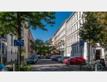 Straßenszene mit mehrstöckigen Altbauhäusern, teils mit verzierten Fassaden und Balkonen, beidseitig entlang einer schmalen Straße. Im Vordergrund parkende Autos, Fahrräder und Straßenschilder, umgeben von Bäumen und Begrünung.