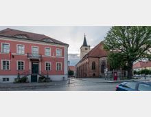 Städtischer Platz mit einem historischen Rathaus in rosafarbenem Außenanstrich und einem Steingebäude mit rotem Ziegeldach sowie Kirchturm im Hintergrund. Links stehen Fahnen am Rathaus, während rechts ein großer Baum und eine Bushaltestelle auf dem gepflasterten Platz zu sehen sind.