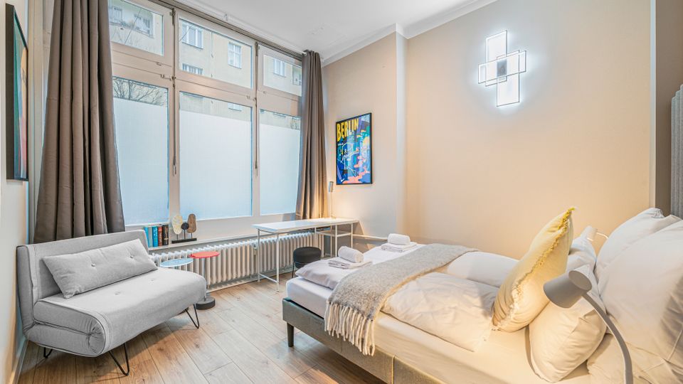 Helles, modern eingerichtetes Schlafzimmer mit großem Bett, kleiner Sitzbank und Schreibtisch vor einer breiten Fensterfront mit dunklen Vorhängen. Auf hellem Holzfußboden stehen ein kleiner Beistelltisch und eine Stehlampe; an der Wand hängt ein Poster und eine geometrische Wandleuchte.