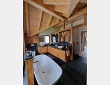 Modernes Badezimmer mit freistehender weißer Badewanne, dunklem Fliesenboden und Holzschränken. Sichtbare Holzdeckenbalken und ein großer Waschtisch mit zwei Waschbecken prägen den Raum.