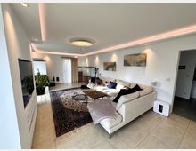 Modernes Wohnzimmer mit heller Einrichtung, bestehend aus einer großen Ecksofa-Landschaft, einem Couchtisch und einem dekorativen Teppich. Der Raum ist mit weißen Wänden, minimalistischer Beleuchtung, großen Fenstern mit Vorhängen und Wandkunst gestaltet, die eine gemütliche Atmosphäre schaffen.