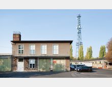 Zweistöckiges Büro- oder Lagergebäude mit einer schlichten Fassade, grün gestrichenen Garagentoren und einem roten Backsteinkamin. Im Hintergrund ein hoher Sendemast und eine Reihe schlanker Bäume.