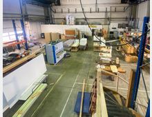 Innenansicht einer Werkstatt mit hohen Decken, Arbeitsstationen und Maschinen. Zu sehen sind Holzmaterialien, Werkbänke, Regale und verschiedene Werkzeuge in einer industriellen Umgebung.