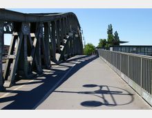 Stählerne Fußgänger- und Straßenbrücke mit Fachwerkstruktur, beleuchtet von Laternen, deren Schatten auf den Gehweg fallen. Rechts daneben verläuft ein Geländer, im Hintergrund sind Bäume und ein Bahnhofsgebäude mit "S"-Symbol sichtbar.