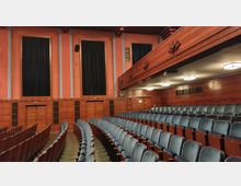 Saal eines Theaters oder Kinos mit gepolsterten Sitzreihen, hölzernen Wandverkleidungen und großen vertikalen schwarzen Vorhängen an den Wänden. Die Decke hat rechteckige Lampen, und ein hölzerner Balkon mit dekorativen Elementen verläuft entlang der rechten Seite.