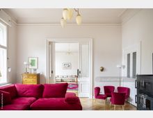 Wohnzimmer mit hellen Wänden und Fischgrätenparkettboden, ausgestattet mit einem roten Ecksofa und passenden kleinen Sesseln. Im Hintergrund ein angrenzender Raum mit einem Sofa, Teppich und einer großen Schiebetür mit Glaselementen.