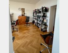 Ein Büro mit Parkettboden und weißen Wänden. Im Raum befinden sich Aktenschränke, Regale mit Aktenordnern, mehrere Bürostühle und ein großer Holzschrank mit Schubladen; an der Wand hängt eine Uhr.