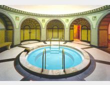 Ein kreisförmiger Innenraum mit einem zentralen Whirlpool, umgeben von dekorativen, mit Mosaiken verzierten Wänden und runden Bögen. Der Raum verfügt über vergoldete Handläufe, symmetrische Muster und eine gleichmäßige Beleuchtung.