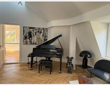 Heller Raum mit schrägen Wänden, in dem ein schwarzer Flügel steht. Auf dem Parkettboden sind eine schwarze moderne Tischlampe, ein dekorativer Bärenfigur sowie ein Sofa platziert, im Hintergrund ein großes abstraktes Gemälde und durch die Tür ist ein weiteres Zimmer mit großen Fenstern sichtbar.