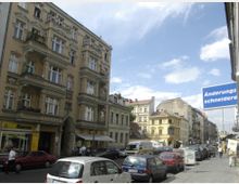 Straßenansicht in einer Stadt mit historischen Wohngebäuden und verzierten Fassaden. Autos parken entlang der Straße, während Geschäfte und Werbeschilder, darunter ein Schild einer Änderungsschneiderei, zu sehen sind; im Hintergrund ist eine U-Bahnstation erkennbar.