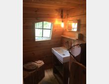 Rustikale Holzkabine mit einem kleinen Badezimmer, bestehend aus einem Waschbecken mit stilisiertem Wasserhahn, einem Spiegel an der Wand und einer Komposttoilette mit Holzdeckel. Ein quadratisches, unterteiltes Fenster und eine hängende Glühbirne sorgen für Licht.