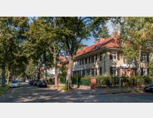 Ecke einer ruhigen Wohnstraße mit Kopfsteinpflaster, gesäumt von Bäumen. Im Vordergrund steht ein zweigeschossiges Wohnhaus mit heller Fassade, braunen Fensterläden und rotem Ziegeldach, umgeben von einem Garten mit grünem Zaun.