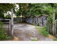 Ein verlassenes Gelände mit mehreren Metallgaragen, deren Türen mit Graffiti bemalt sind. Im Hintergrund stehen Bäume und ein Wohngebäude, während ein verrostetes Gittertor im Vordergrund sichtbar ist.