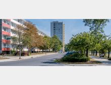 Straßenszene mit einem Wohngebiet: Links im Bild mehrstöckige Wohngebäude mit Balkonen und roten Akzenten, rechts eine mit Bäumen gesäumte Gehweg- und Parkfläche, auf der Autos parken und eine Person auf einer Bank sitzt. Im Hintergrund ein weiteres, höheres Hochhaus.