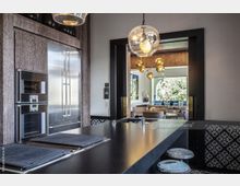 Moderne Küche mit dunkler Arbeitsfläche, integriertem Herd und Einbaugeräten aus Edelstahl. Im Hintergrund ist ein offener Wohnbereich mit großen Fenstern, geschmückt mit goldenen Hängeleuchten und Sitzmöbeln, zu sehen.