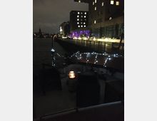 Nachtaufnahme einer Uferpromenade mit modernen Gebäuden, die von verschiedenen Lichtquellen und violetter Beleuchtung erhellt werden. Im Vordergrund sind ein Boot mit kleinen Lichterketten und eine beleuchtete Laterne zu sehen, dahinter fließt ein Fluss.