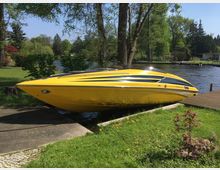 Gelbes Motorboot auf einer Slipanlage am Ufer eines Sees, umgeben von grünem Rasen, Bäumen und kleinen Gebäuden im Hintergrund.