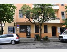 Fassade eines Wohn- und Geschäftsgebäudes in einer Stadt mit beigefarbenem Putz und Balkonen. Im Vordergrund stehen ein Baum, ein geparktes Auto, ein Fahrrad und ein Lieferwagen, während an den geschlossenen Rollläden Graffiti sichtbar ist.