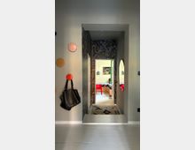 Narrow interior hallway with gray walls, a patterned feature wall around a doorway, and a rug leading into a bright dining area with red chairs. Round wall hooks and a hanging bag are on the left wall, and an oval wall mirror is mounted on the right.