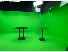 Zwei schwarze Tische stehen in einem Raum mit vollständig grünen Wänden, Boden und Decke, eingerichtet als Greenscreen-Studio. Oben hängen Beleuchtungselemente und schwarze Stoffbahnen.