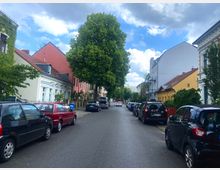 Wohnstraße mit geparkten Autos auf beiden Seiten, flankiert von bunten Wohnhäusern mit roten und gelben Fassaden sowie einem großen Baum in der Mitte der Straße. Der Himmel ist teils bewölkt, und die Straße wirkt ruhig.