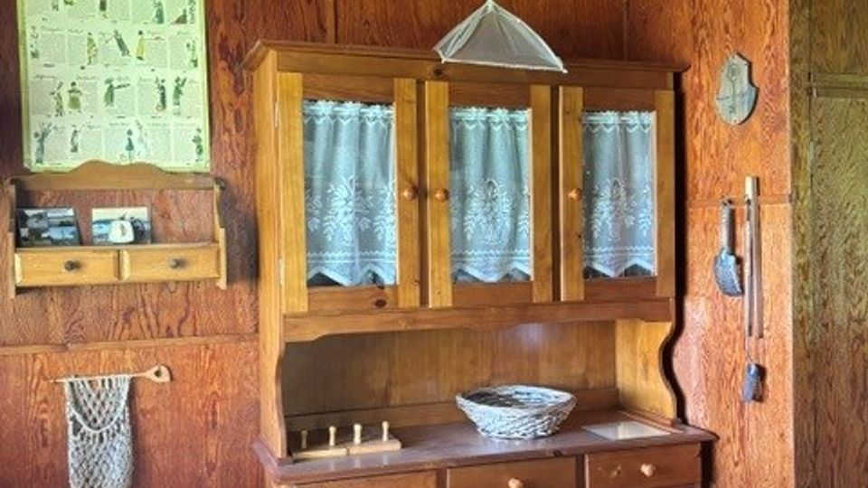 Holzvertäfelter Innenraum mit einem traditionellen Holz-Buffetschrank mit Glasfront und Spitzenvorhängen, darüber eine Hängelampe und ein kleiner Deckenventilator. An der Wand hängen ein Poster und ein kleines Regal, daneben befinden sich Haken und Küchenutensilien neben einer Tür.