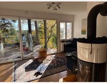 Wohnzimmer mit Holzfußboden und einem Teppich, einem Kaminofen im Vordergrund und einem blauen Sofa im Hintergrund. Große Glasfenster bieten Blick auf eine Terrasse mit Holzgeländer und einen herbstlichen bewaldeten Außenbereich mit einem See im Hintergrund.