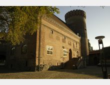 Historischer Backsteinbau mit großem Turm und Spitzbogenportal, umgeben von Kopfsteinpflaster. Im Vordergrund ein Baum und eine Straßenlaterne, rechts eine Brücke.