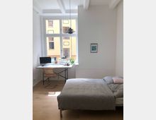 Helles Schlafzimmer mit einem großen Fenster, das auf ein gelbes Gebäude blickt. Im Raum befinden sich ein Bett mit grauer Bettwäsche, ein Schreibtisch mit Holzplatte und schwarzem Gestell, ein Stuhl sowie einige Pflanzen auf dem Tisch; eine Deckenleuchte und ein kleines Wandbild runden die Einrichtung ab.