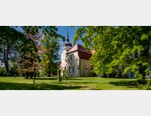 Kirche mit rotem Satteldach und hohem Turm mit dunkler Spitze, umgeben von grünen, dicht belaubten Bäumen auf einer Wiese. Im Vordergrund stehen kleine Denkmäler und Büsche, die das Bild bereichern.