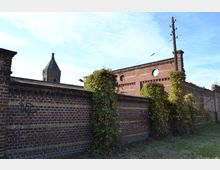 Backsteinmauer mit gelblichen Zierstreifen, überwuchert von Pflanzen, im Vordergrund. Dahinter Ruinen eines Backsteingebäudes mit runden Fensteröffnungen, und ein Kirchturm mit spitzem Dach ist im Hintergrund sichtbar.