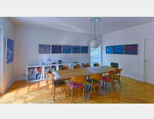 Heller Besprechungsraum mit einem großen Holztisch, umgeben von Stühlen mit gemusterten Polstern. An den weißen Wänden hängen abstrakte Gemälde, und ein niedriger Bücherregal steht an der Rückwand unter den Bildern. Eine große, kugelförmige Deckenleuchte hängt über dem Tisch.