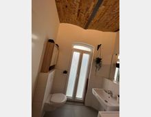 Kleines Badezimmer mit weißen Fliesen, an der linken Wand ein hängendes WC, rechts ein Waschbecken mit Spiegel. Das Fenster in der Mitte hat eine Milchglasverkleidung, und die Decke besteht aus freiliegendem Ziegelmauerwerk mit Metallträgern.