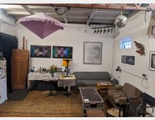 Ein gemütlicher Raum mit schlichtem weißen Wänden, ausgestattet mit Sofas, einem kleinen Tisch und einer Theke mit Blumen und Getränken. Dekorationselemente wie eine Violette Hängelampe, eine Diskokugel, moderne Wandbilder und ein ausgestopfter Vogel bringen einen kreativen und unkonventionellen Stil.