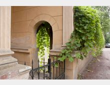 Ansicht einer historischen Sandsteinmauer mit Säulen und einem Rundbogen, durch den Blick auf einen grünen Garten mit hängendem Weinlaub fällt. Ein schmiedeeisernes Tor befindet sich im Vordergrund, und ein Weg führt rechts an der Mauer entlang.