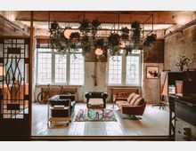 Industriell eingerichteter Raum mit Ziegelwänden, großen Sprossenfenstern und freiliegender Decke; im Vordergrund stehen Sofas, ein Couchtisch und ein Teppich, darüber hängen Pflanzen und Pendelleuchten, im Hintergrund ein Fahrrad vor einem Heizkörper.