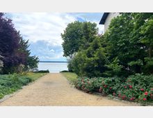 Gartenweg mit Kiesbelag, flankiert von roten Rosensträuchern und dichten Bäumen, der zu einem See führt; im Hintergrund ist eine Uferbarriere und bewölkter Himmel sichtbar. Links und rechts des Weges befindet sich üppige Vegetation, rechts ragt ein Teil eines Gebäudes ins Bild.