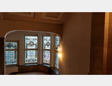 Ein Innenraum mit verzierten Buntglasfenstern in einem Erker, die detaillierte Wappen und Muster zeigen. Die Wände sind schlicht gehalten, mit einem modernen Wandlicht an der Seite, und die Decke hat eingelassene Kassettenelemente.