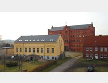 Gelbes, einstöckiges Gebäude im neoklassizistischen Stil mit Satteldach und großen Fenstern, daneben ein großes rotes Backsteingebäude im industriellen Stil mit mehreren Etagen; umgeben von einer Wiese und einem Kiesweg, im Hintergrund offene Landschaft.