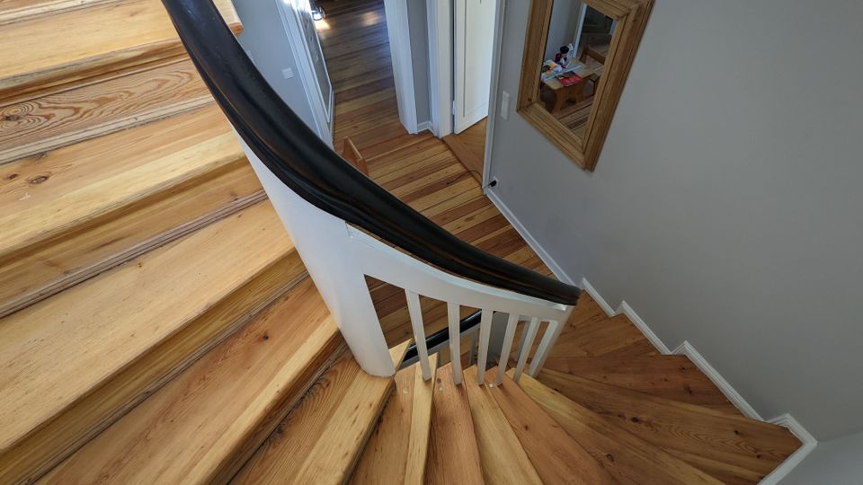 Blick von oben auf ein geschwungenes Holztreppenhaus mit hellen Stufen, weißem Geländer und dunklem Handlauf. Unten führt ein Flur mit Holzboden zu mehreren Türen; an der Wand hängt ein rechteckiger Spiegel mit Holzrahmen.
