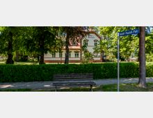 Blick auf ein historisches Kurgebäude mit weißer Fassade und rotem Ziegeldekor, umgeben von hohen Bäumen und Grünanlagen. Im Vordergrund stehen eine Parkbank, eine Hecke entlang des Gehwegs und ein Straßenschild „Rudolf-Poschich-Platz“.
