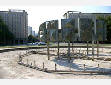 Ein im Freien stehender, nicht in Betrieb befindlicher Brunnen mit metallischen, geometrisch gestalteten Skulpturen auf Säulen, umgeben von einer runden, betonierten Fläche mit Rohrleitungen. Im Hintergrund befinden sich große, mehrstöckige Wohngebäude im architektonischen Stil der Nachkriegszeit sowie eine Straße mit wenig Verkehr.