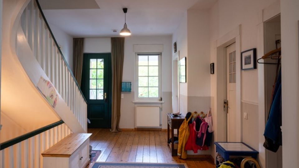 Heller Hausflur mit Holzfußboden, Treppe mit weißem Geländer und grünem Handlauf sowie einer grünen Eingangstür. Neben einem Fenster mit Vorhängen stehen eine kleine Bank mit Garderobe, ein Tisch und eine Deckenleuchte; rechts sind Innentüren und Jacken an Haken.