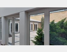 Innenhof mit großen Glasfenstern, die in einen gepflasterten Außenbereich führen, umgeben von gelben Backsteinwänden; im Vordergrund befinden sich grüne Sträucher.