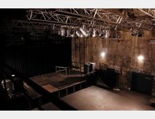 Ein leerer Veranstaltungsraum mit einer erhöhten Bühne aus dunklem Material, umgeben von schwarzen Vorhängen. Die Wände sind aus Beton und teils mit Graffiti bedeckt, während von der Decke ein Gitterrahmen mit zahlreichen Scheinwerfern hängt.