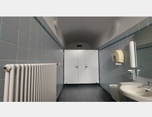 Öffentliche Toilettenanlage mit zwei weißen Kabinentüren an der Rückwand, hellblauen Wandfliesen, grauem Bodenfliesenbelag, einem Waschbecken mit Seifenspender, einem weißen Heizkörper und einem Papierhandtuchspender.