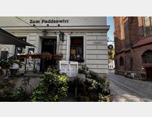 Fassade eines historischen Gebäudes mit dem Schriftzug „Zum Paddenwirt“ in einer gepflasterten Gasse. Vor der Tür stehen Pflanzen und Blumenkästen, neben dem Eingang hängt ein Menüschaukasten, und im Hintergrund ist ein Backsteinbau mit gotischen Fenstern zu sehen.