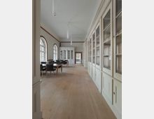 Heller Raum mit historischen Einbauschränken aus Holz und verglasten Türen, dazu eine Reihe von Tischen mit schwarzen Stühlen. Große, bogenförmige Fenster lassen Tageslicht herein, und der Boden besteht aus hellem Holz.