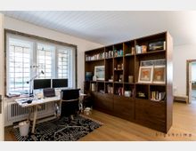 Arbeitszimmer mit einem Schreibtisch vor drei Fenstern mit dekorativen Gittern, einem drehbaren Bürostuhl und zwei Monitoren. Rechts steht ein großer, offener Holzregal mit Büchern, Dekorationsgegenständen und gerahmten Bildern.