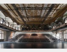 Großer, leerer Veranstaltungssaal mit einer zentralen Tribüne aus gestaffelten Sitzreihen und offenem Bühnenboden im Vordergrund. Hohe Decke mit abblätterndem Putz, sichtbarer Traversenkonstruktion und umlaufenden Galerien sowie großen Rundbogenfenstern entlang der Seitenwände.